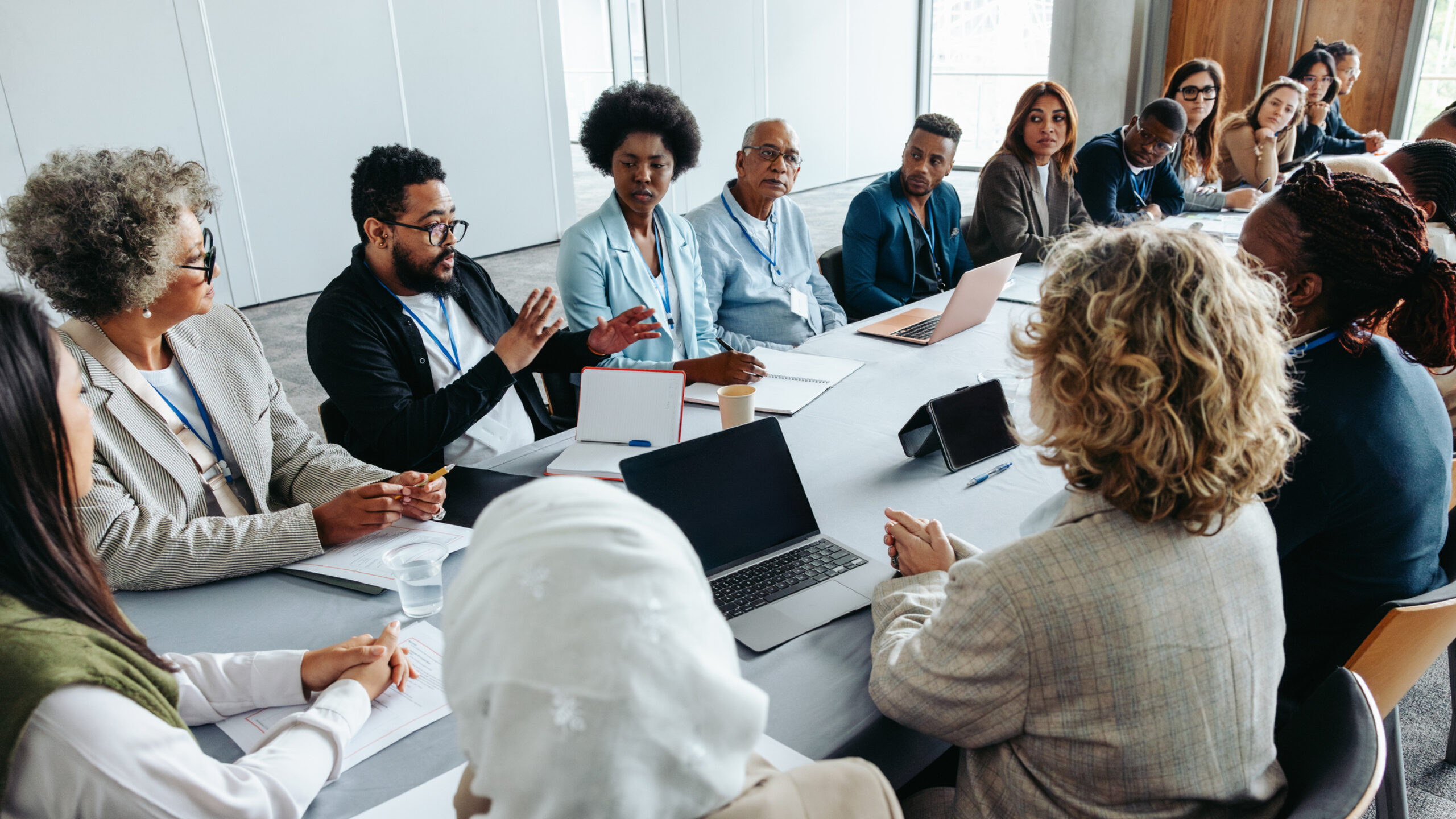 A diverse group of professionals sit around a conference table in a modern meeting room, engaged in discussion during a collaborative meeting, with laptops, notebooks, and coffee cups on the table.