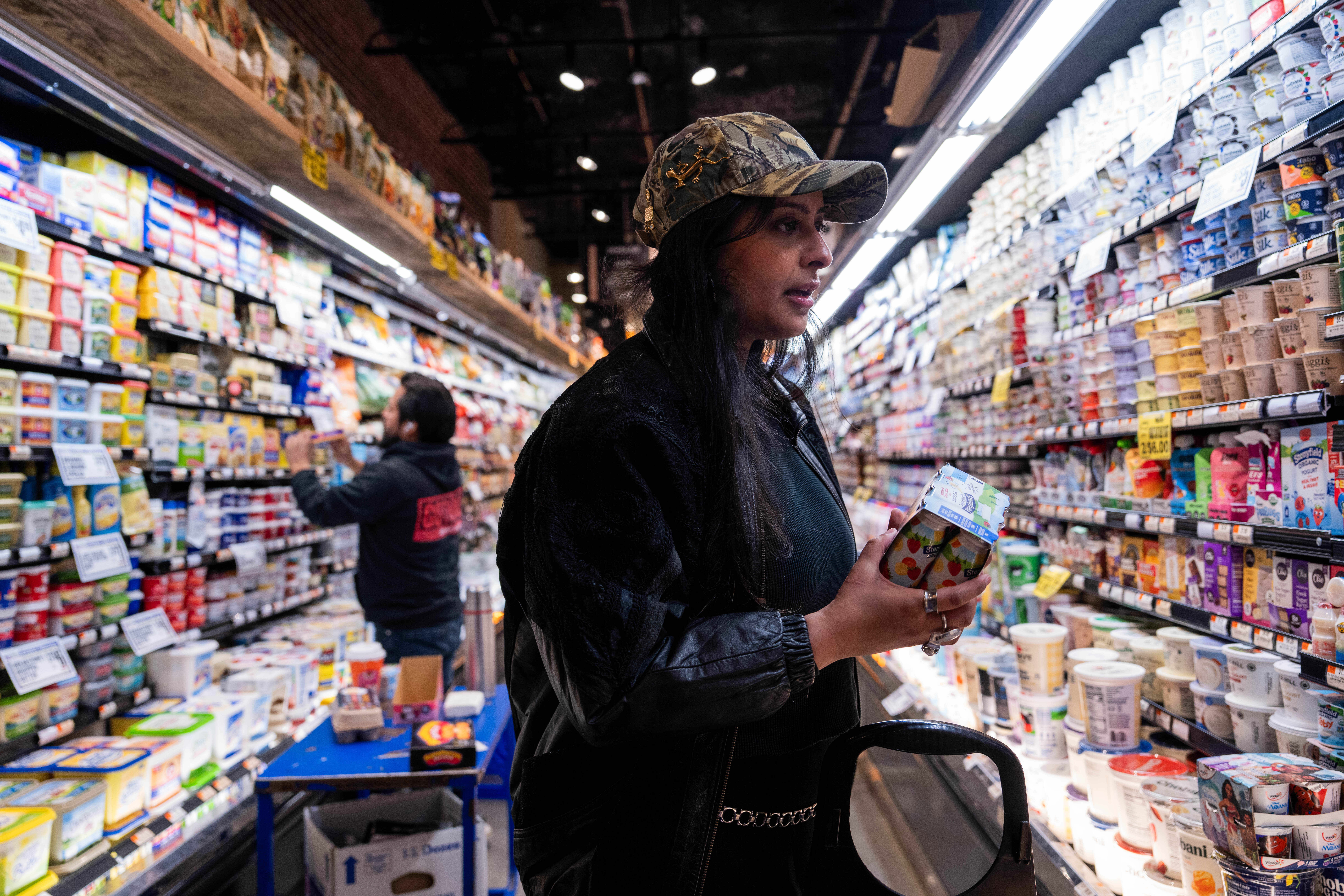 A woman in a camouflage hat holds a pack of yogurt in a brightly lit supermarket dairy aisle, surrounded by shelves of various dairy products.
