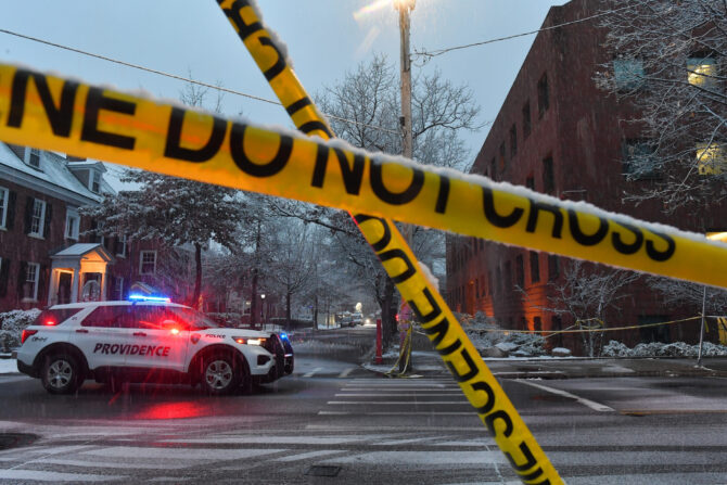 A Providence Police SUV with flashing red and blue lights is parked on a snowy city street at night, behind yellow crime scene tape. Buildings and bare trees are visible in the background.
