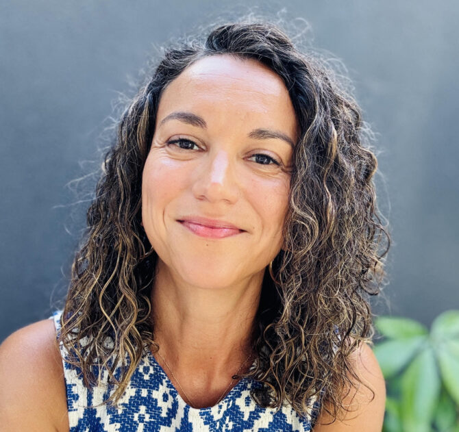 A smiling woman with curly dark hair and a blue and white patterned top looks directly at the camera against a dark gray background.