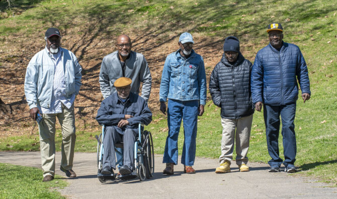 Six older Black men, five standing and one in a wheelchair, pose together on a paved path in a park.
