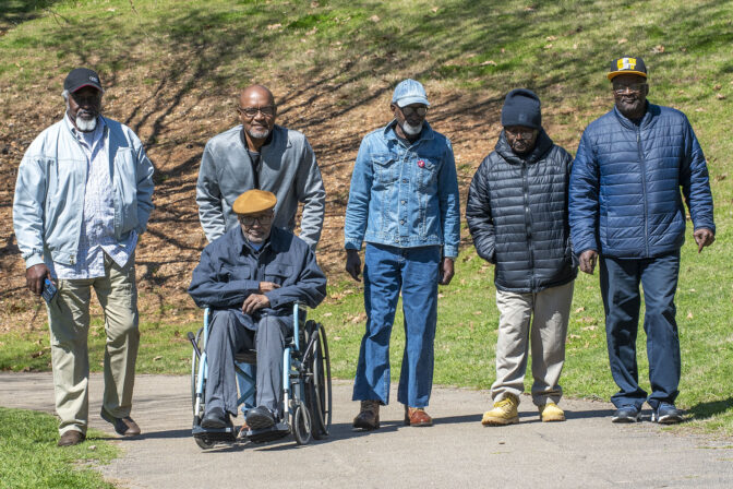 Six older Black men, five standing and one in a wheelchair, pose together on a paved path in a park.
