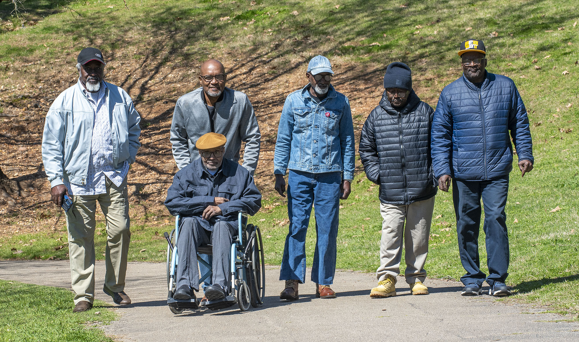 Six older Black men, five standing and one in a wheelchair, pose together on a paved path in a park.