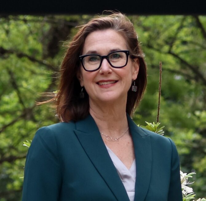 A smiling woman with reddish-brown hair and black-framed glasses wears a dark teal blazer against a blurred green foliage background.
