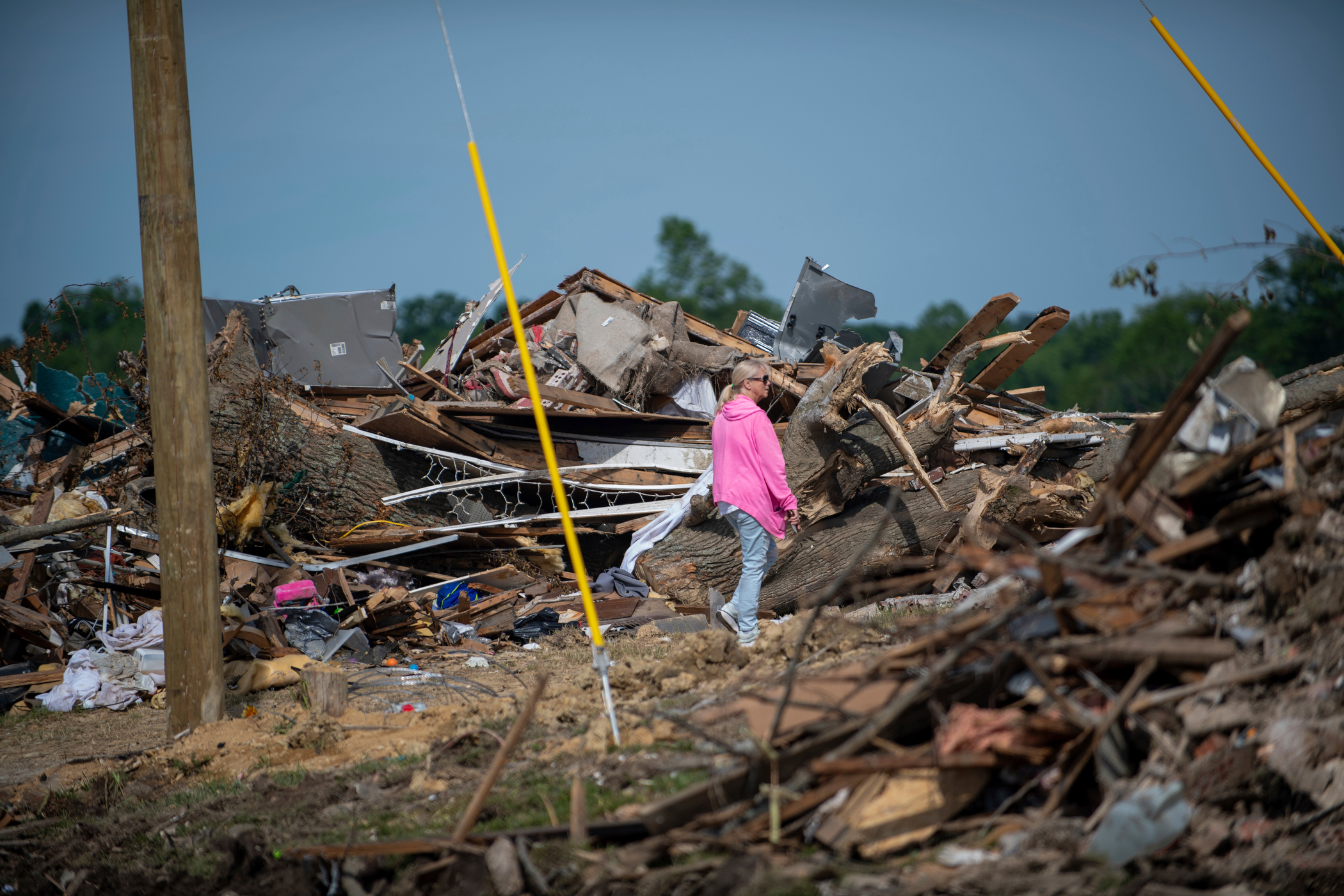 A person in a pink hoodie walks through a vast field of tornado or storm debris, including splintered wood, tree trunks, and household items, under a blue sky.
