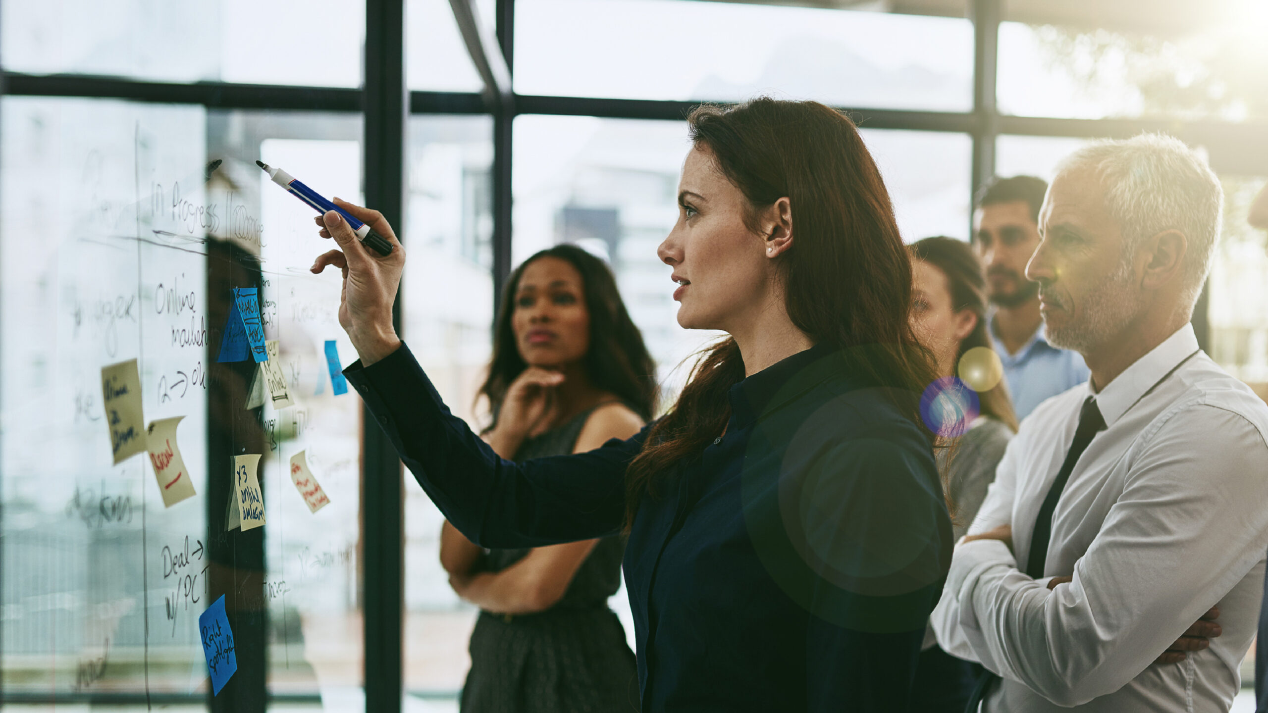 A woman writes on a glass whiteboard covered with notes and diagrams while a small group of colleagues stands behind her, watching and collaborating in a modern office setting.