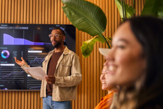 A professional presents data and analytics on a large digital screen during a meeting, gesturing with a document in hand while colleagues listen in a modern, plant-filled workspace.