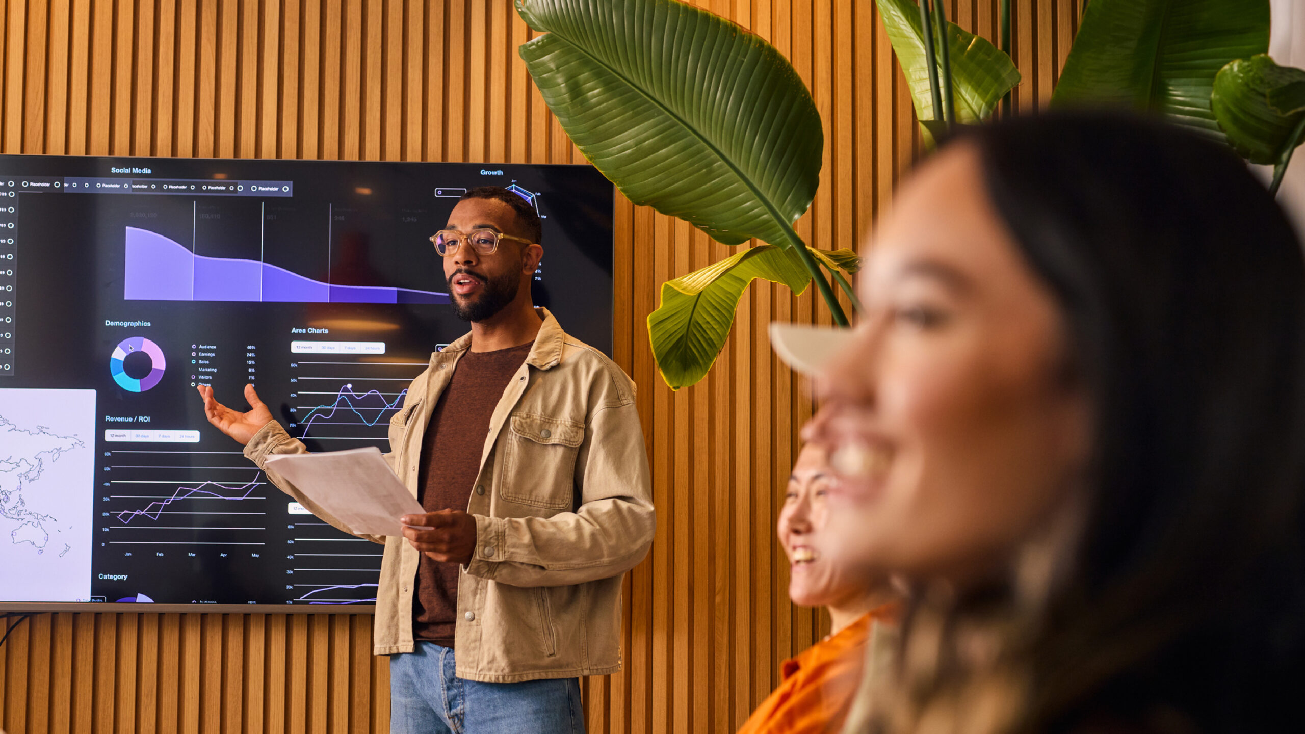 A professional presents data and analytics on a large digital screen during a meeting, gesturing with a document in hand while colleagues listen in a modern, plant-filled workspace.