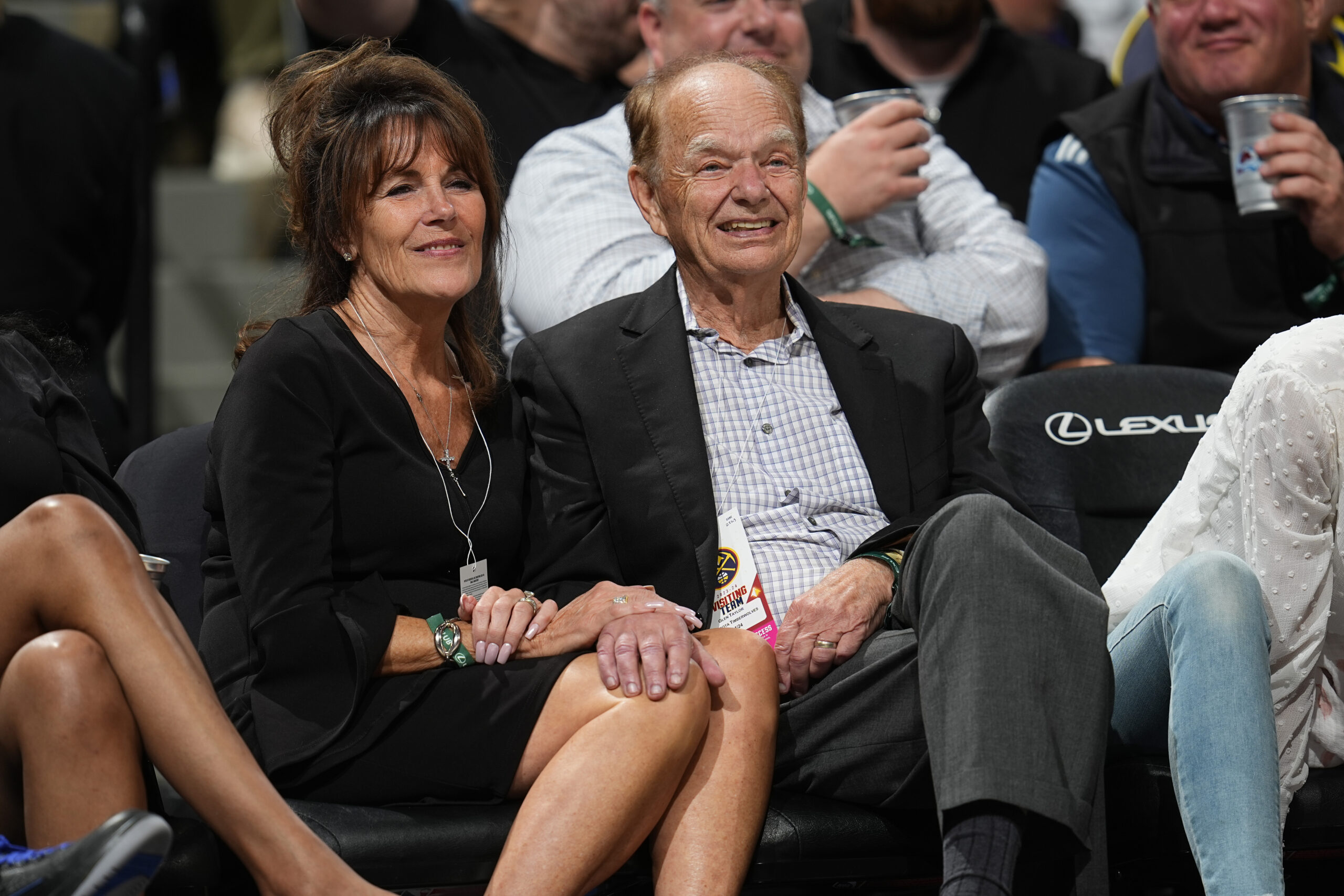 An older man and a middle-aged woman, Glen and Becky Taylor, smiling and holding hands at a Minnesota Timberwolves game. The man wears a blazer and a 'Visiting Team' pass.