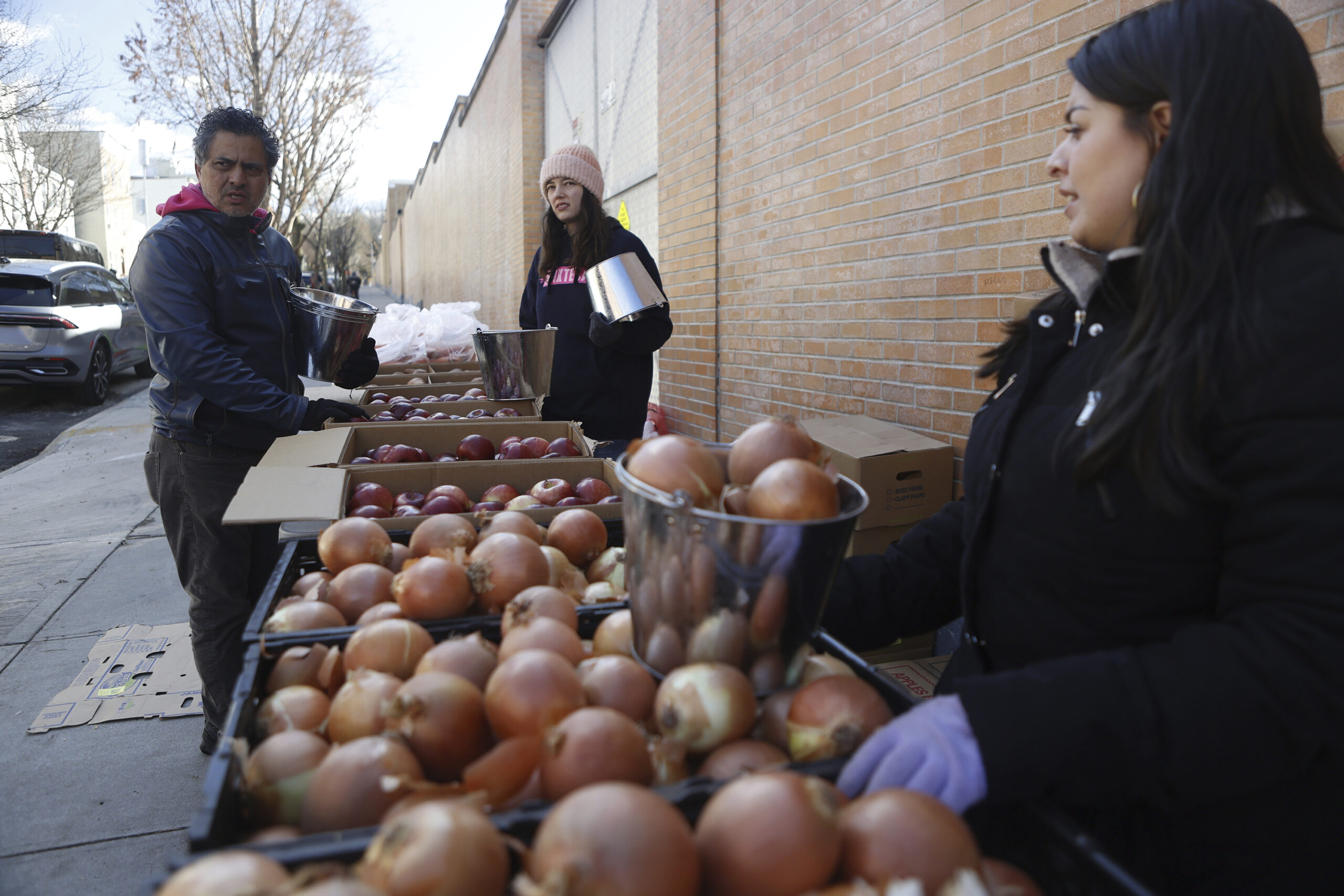 Three people, two women and one man, organize crates of red apples and brown onions on a sidewalk next to a brick wall. They are holding metal buckets and appear to be distributing food.