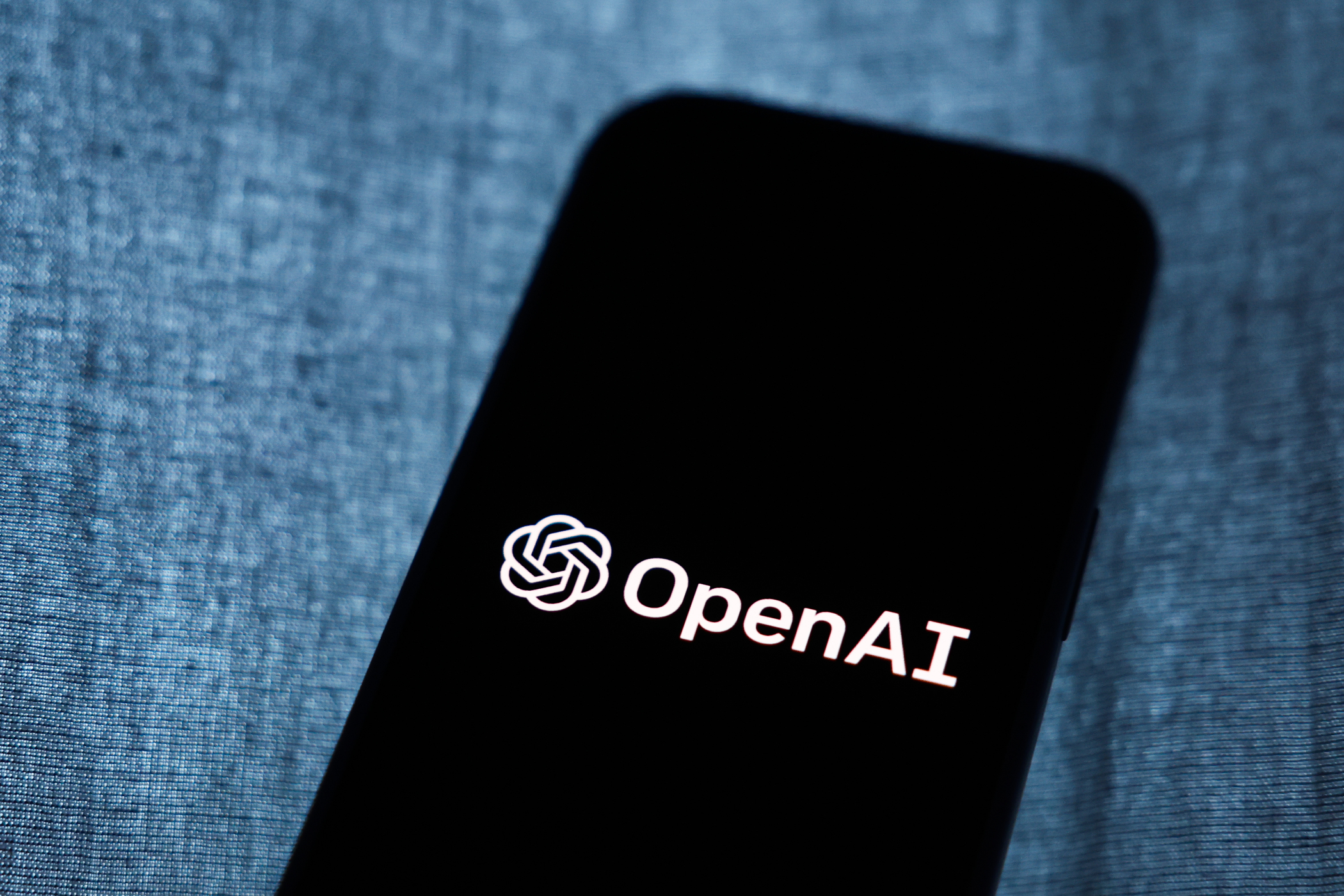 A black smartphone screen displaying the white OpenAI logo and text on a dark background, resting on a blurred blue-grey textured fabric.