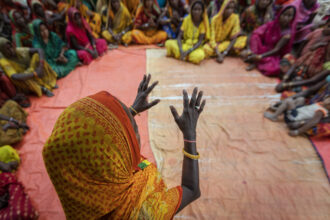 Overhead view of a woman in a yellow and orange sari with raised hands, facing a circle of other women in colorful traditional clothing seated on an orange tarp.