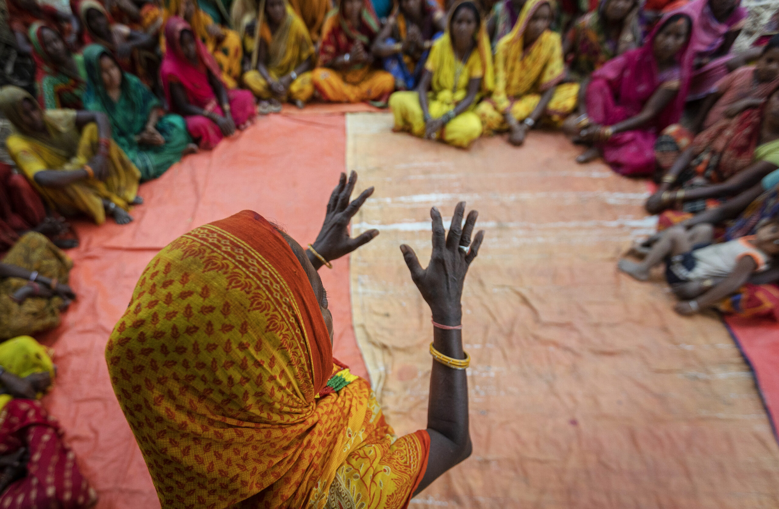 Overhead view of a woman in a yellow and orange sari with raised hands, facing a circle of other women in colorful traditional clothing seated on an orange tarp.