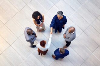 Overhead view of six diverse business professionals forming a circle on a tiled floor, with two women shaking hands in the center.