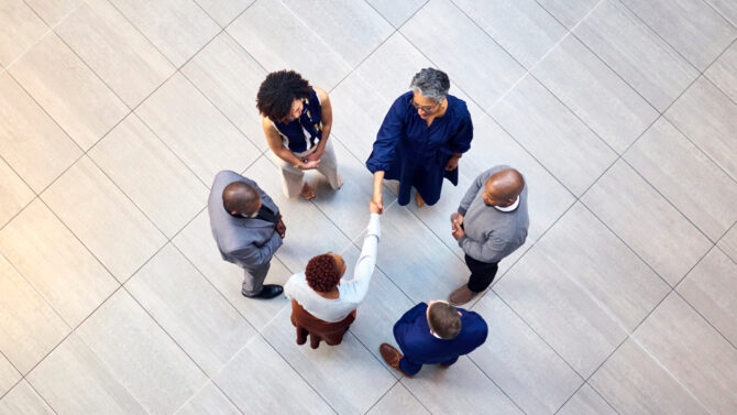 Overhead view of six diverse business professionals forming a circle on a tiled floor, with two women shaking hands in the center.