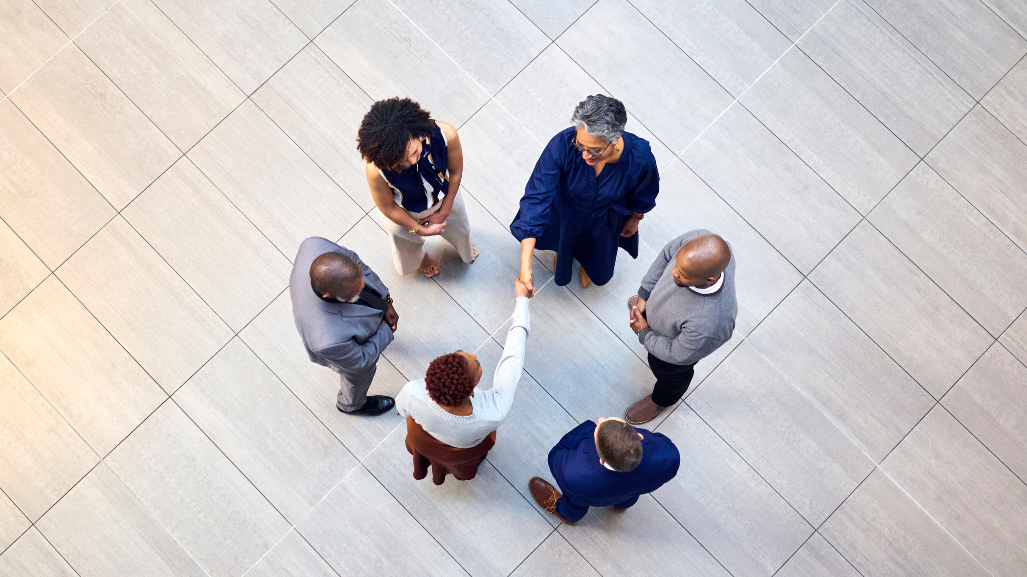 Overhead view of six diverse business professionals forming a circle on a tiled floor, with two women shaking hands in the center.