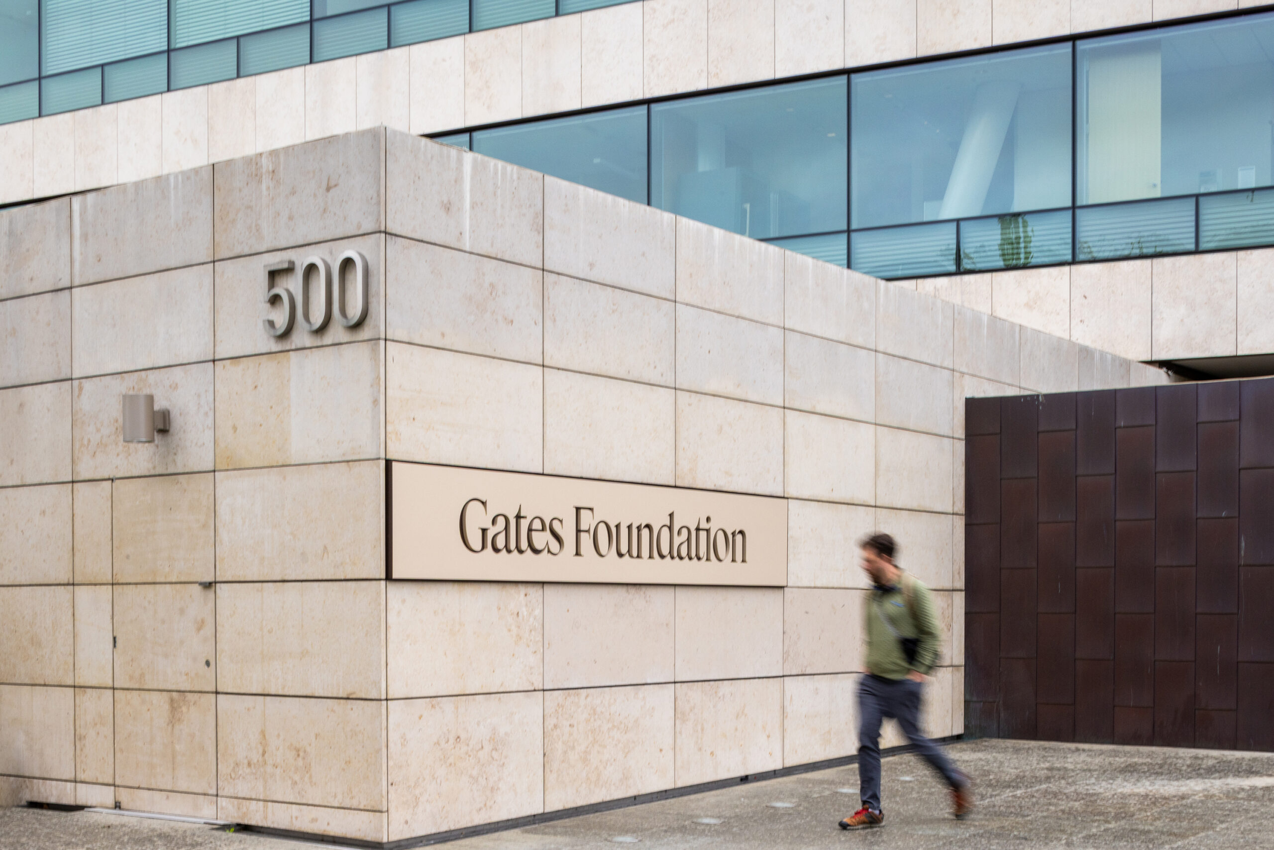 Exterior view of the Gates Foundation building, showing its light beige tiled facade, large reflective windows, and a prominent sign reading 'Gates Foundation'. The building number '500' is also visible. A person in motion blur walks past on the sidewalk.