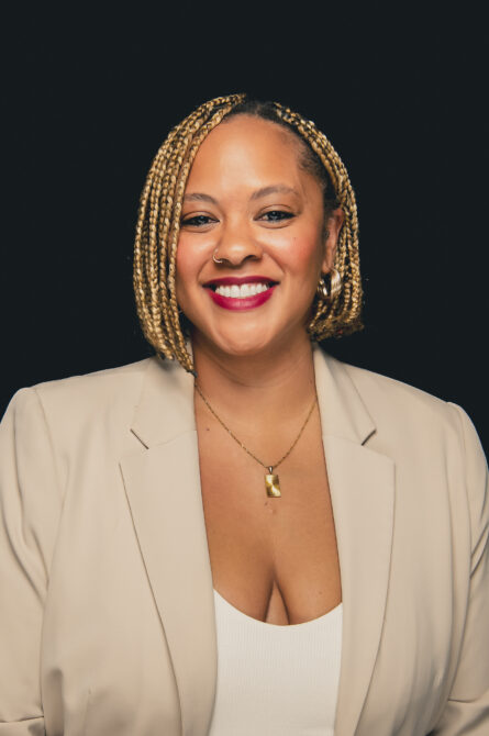 A smiling woman with a nose ring, and red lipstick, wearing a beige blazer and gold necklace against a black background.