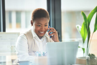 Alt text: A professional woman sits at a desk in a bright office, smiling while talking on the phone and working on a laptop, with a coffee cup and green plant nearby.