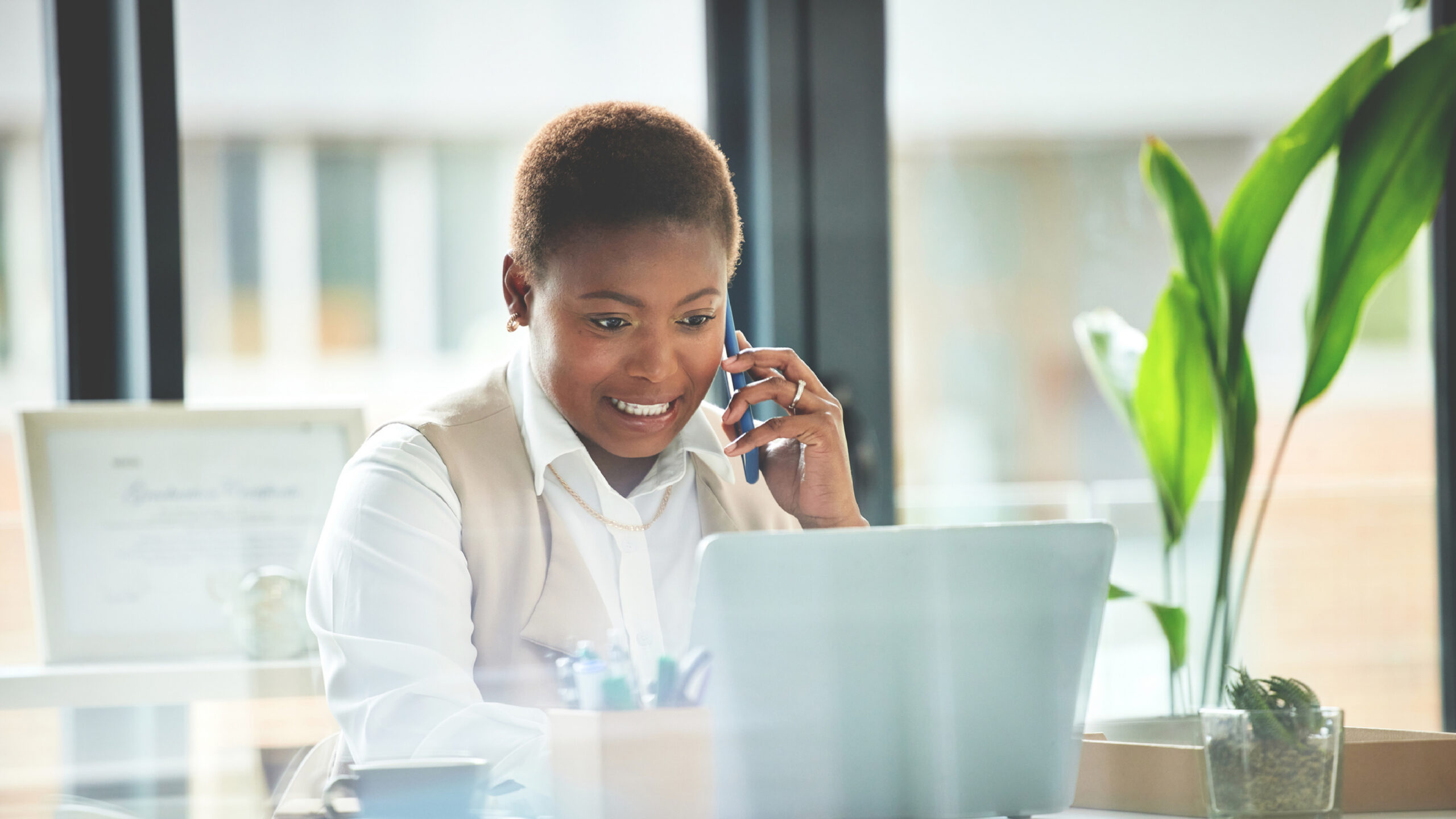 Alt text: A professional woman sits at a desk in a bright office, smiling while talking on the phone and working on a laptop, with a coffee cup and green plant nearby.
