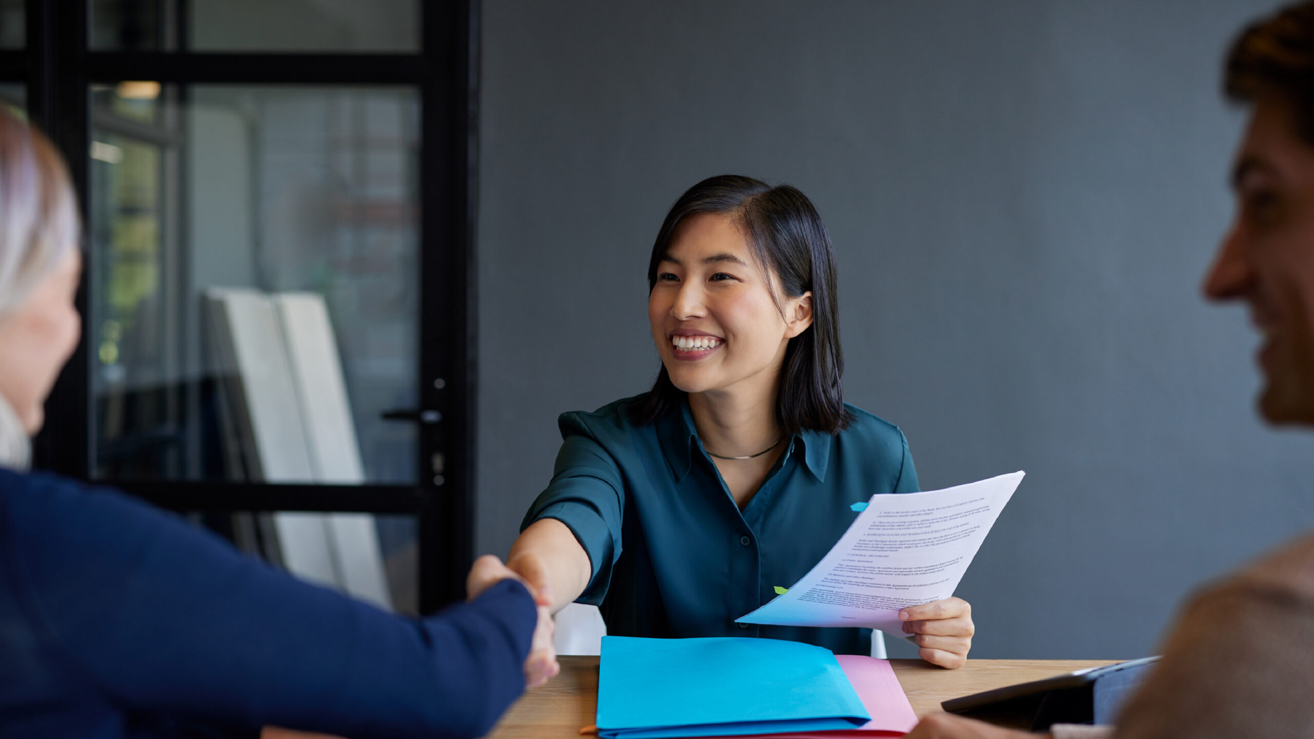 A smiling professional woman sits at a table, shaking hands with a colleague while holding a document, as another person looks on during a meeting.