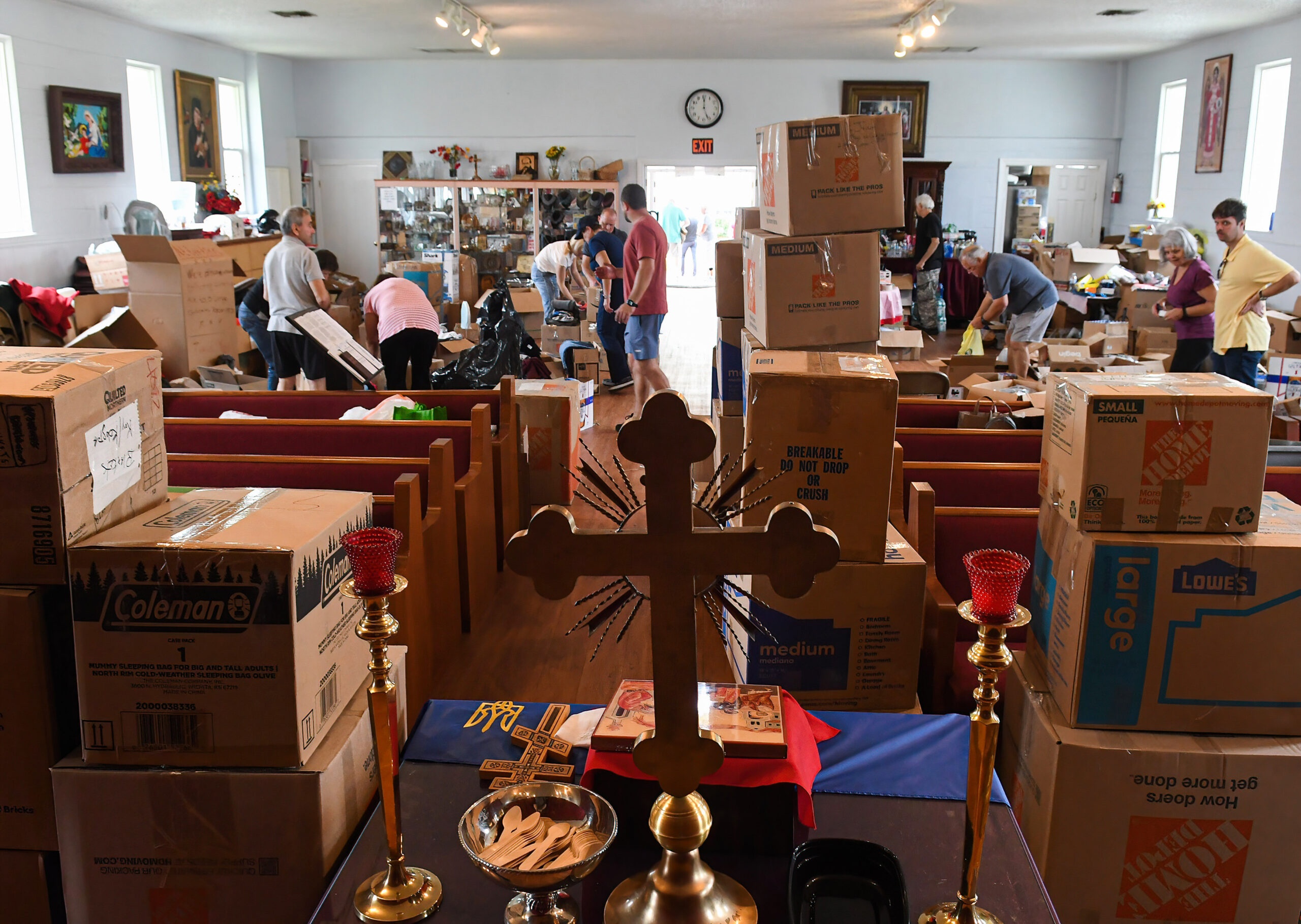 Inside a church hall, volunteers sort numerous cardboard boxes for aid. In the foreground, a wooden cross, candlesticks, and a Ukrainian flag are visible on an altar.